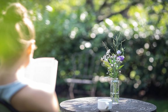 Person sitzt im Garten auf einem Stuhl und liest ein Buch, Tisch mit Tasse und Blumen, Retreat Brandenburg